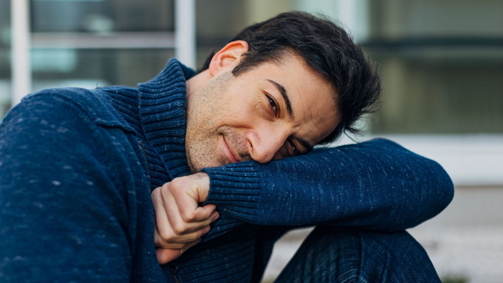 Man in blue sweater resting chin on arm, smiling softly.
